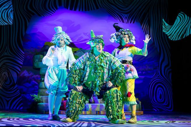 Three actors on stage dressed as the characters in The Baddies, with a dark purple background.