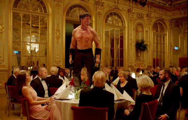 image shows man with no top on stood on a tabel in a formal room surrounded by people wearing formal attire