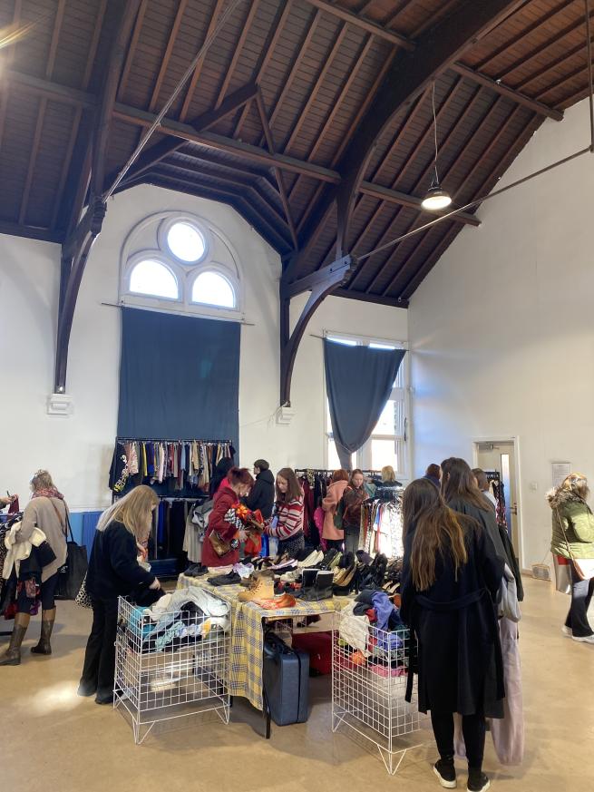 A group of people rummaging through white baskets full of clothes in a community centre hall with high wooden ceilings.