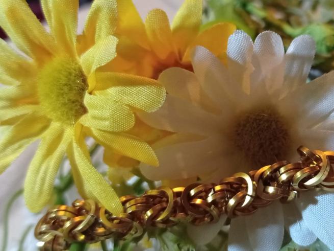 closs up photo of a gold coloured cahinmaille bracelet accross some plastic plants