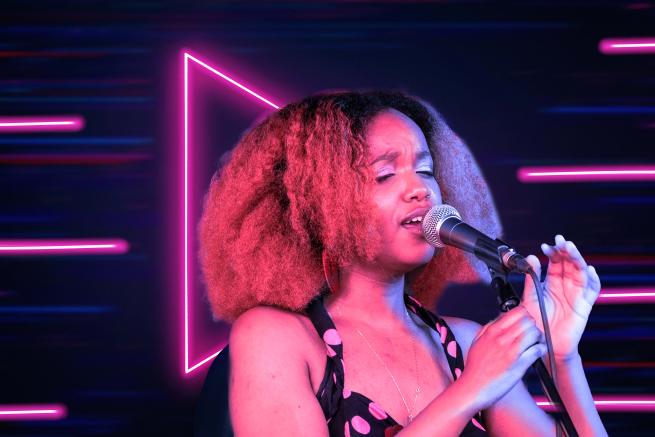 A young lady, in mid-close up, holds a microphone stand with hands almost clasped together, as she sings into the microphone with eyes closed. Her frizzy hair is coloured red and brushed to full volume. Her vintage polka-dotted halter-neck dress contrasts with the background's futuristic neon-pink lines and dark blue-purple background.    