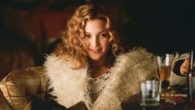 A young woman with blonde curly hair and a large white fur coat sits at a table next to two drinks glasses