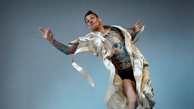 A person with tattoos mid-air as they jump on a bed. They are wearing a dressing gown. 