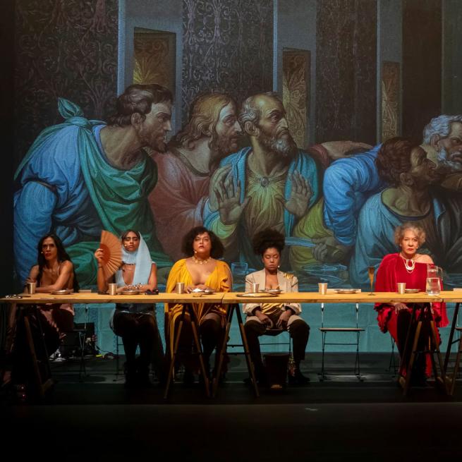 A reinterpretation of ‘The Last Supper’ painting with people gathered around a table. In the background, the original artwork. 