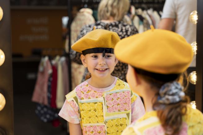 A girl in a yellow hat smiles at her reflection in a mirror