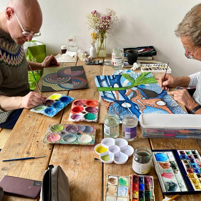 Two men sit at a table with lots of art materials on the table. They look relaxed and content.