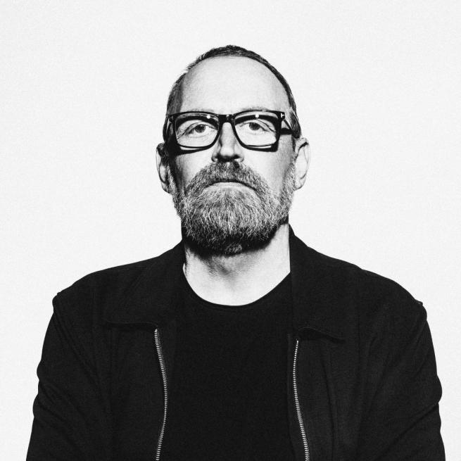 Black and white headshot image of Boo Hewerdine looking straight on at the camera wearing a bomber jacket, tshirt and glasses.