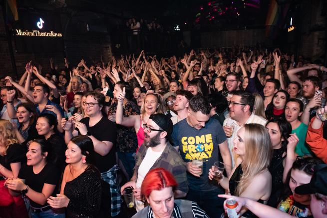 A large, excited crowd of musical theatre fans cheering, dancing, and singing along in a packed nightclub. Many are smiling, clapping, and wearing themed outfits, including a 'Come From Away' t-shirt. The atmosphere is lively, joyful, and full of energy, with colourful lighting and a sign reading 'Musical Theatre Raves' in the background