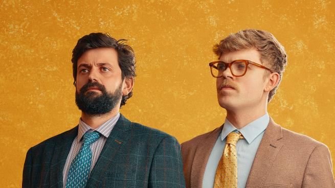 Two people in colorful suits with serious expressions. One has blond hair and red glasses, the other brown hair and a beard.