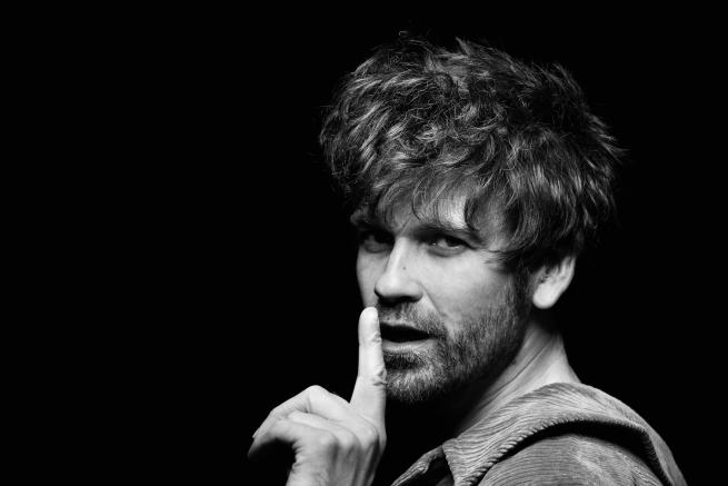 Black and white photograph of Fyfe Dangerfield (white male in his early 40s with brown hair) with his fingers to his lips.