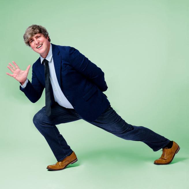 Glenn Moore in a blue suit leaning to the right waving his right hand.