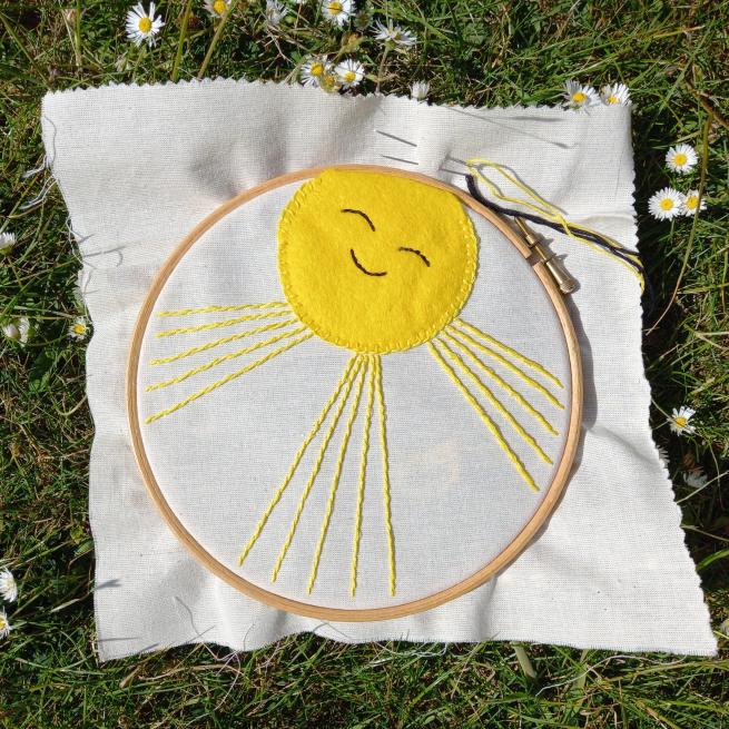 An embroidery hoop with some plain coloured fabric which has been embroidered with a yellow sunshine which has a smile and closed eyes.