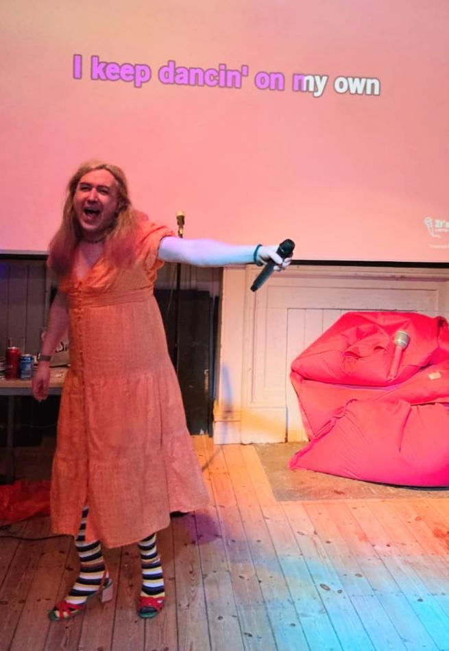 A photo of your host, Quinn a non-binary person with long hair wearing a peach coloured dress, black and white striped tights and red heels singing in front of a karaoke projection screen