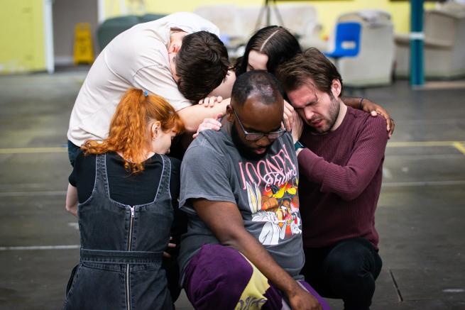 Five individuals with their bodies interconnected through movement, laying on top of each other’s body weight.