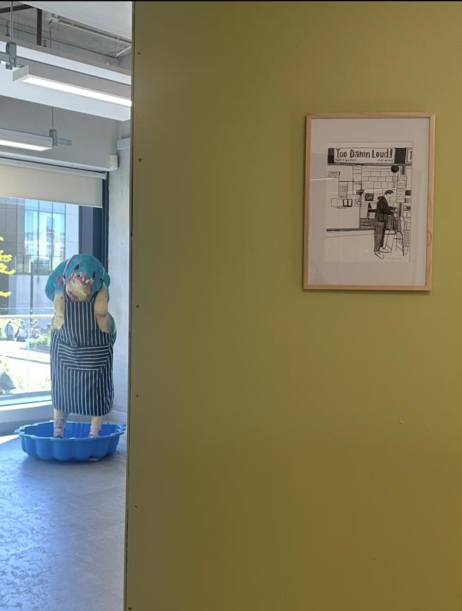 A gallery space with a green wall that has a framed drawing of a music shop and a sculpture of a creature in the space to the left. 
