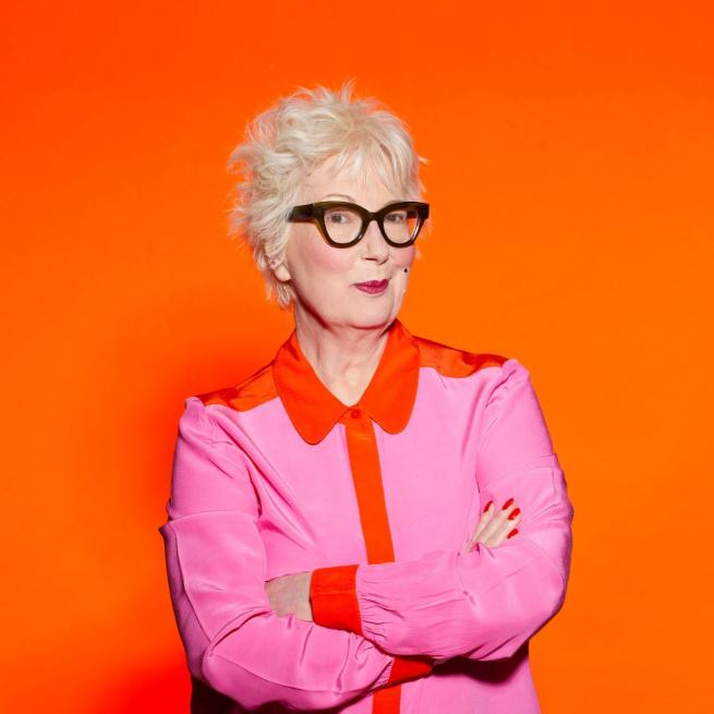 Jenny Eclair with her arms crossed wearing a pink and orange shirt and her signature black glasses against an orange background.