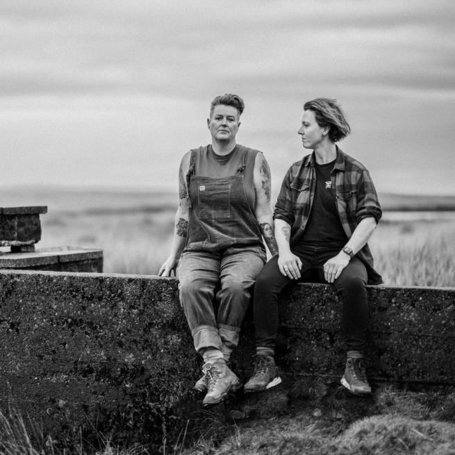 Black and white image of Belinda O'Hooley and Heidi Todow sat on a concrete wall next to a small footbridge with long grass in the background.