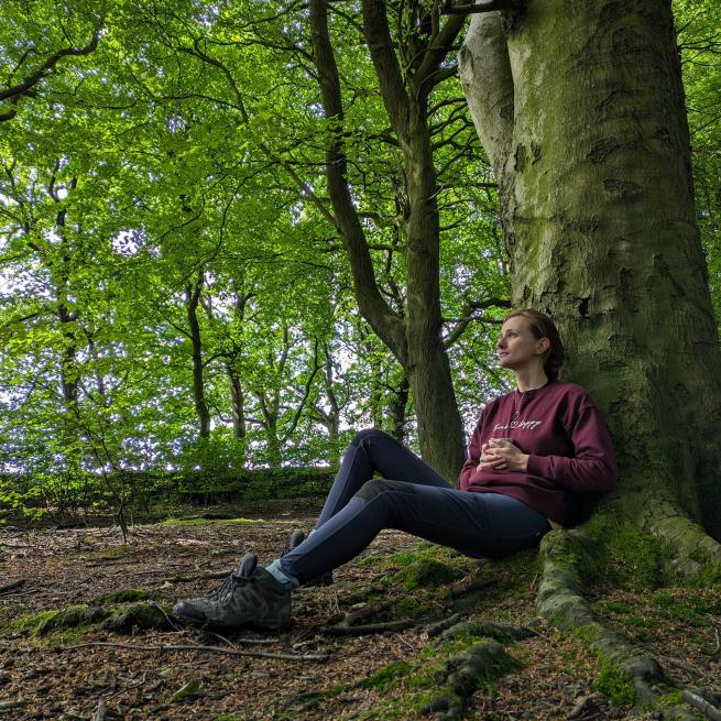 A lady sitting resting against a tree looking pensive with a mug of tea, in a green leafy woodland
