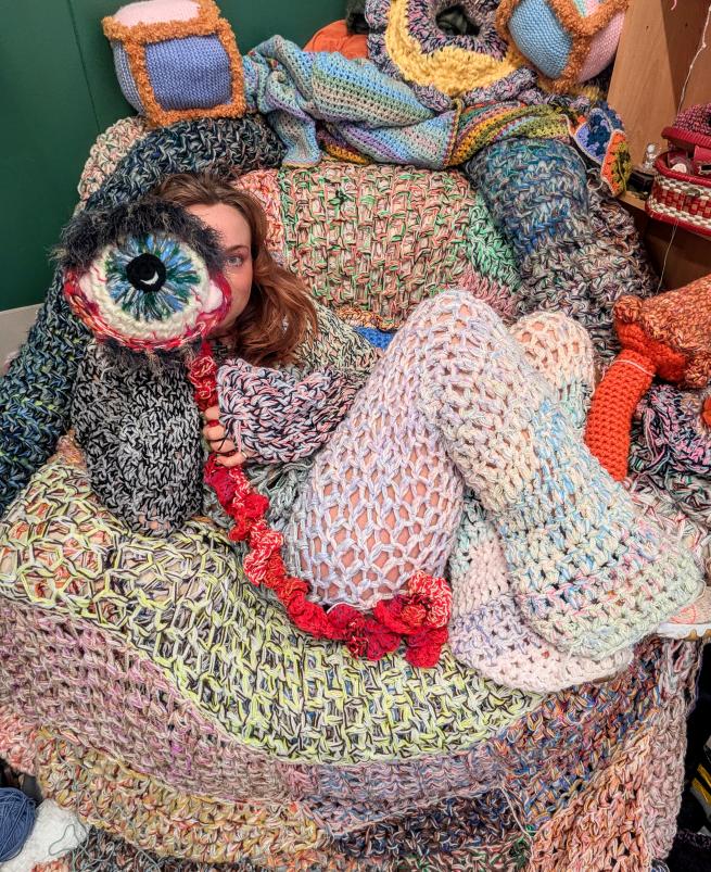 A woman sits on a crochet covered armchair, wearing a crochet costume and holding a crochet eyeball to her face