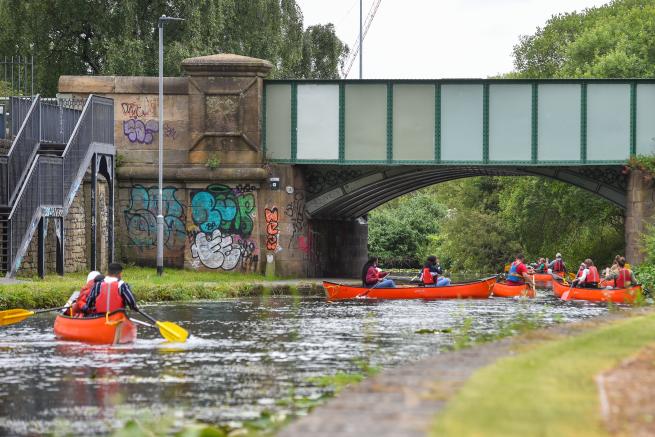 Image of people on 5 canoes on a canal, under a bridge