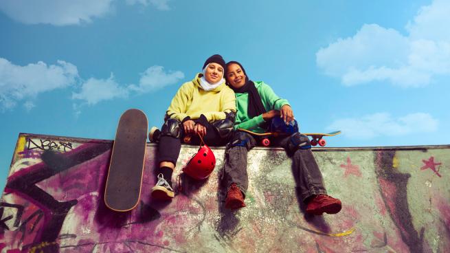 Two girls sit leaning on each other at the top of a skateboard ramp. They hold skateboards and are wearing protective skateboarding gear.