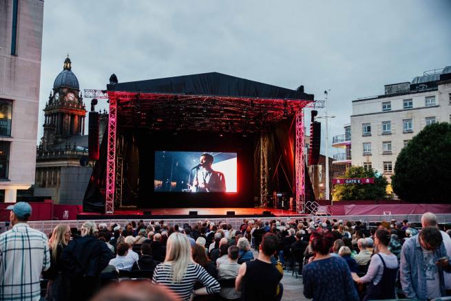 A crowd of people stand in front of a large concert tent with an illuminated screen inside it. 