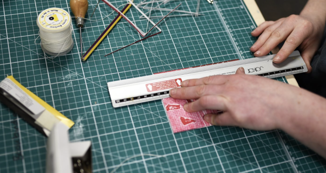 A handbound book being finished with a metal ruler, surrounded by various bookbinding equipment.