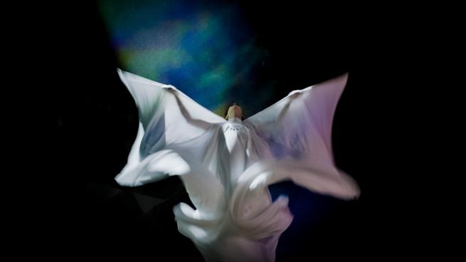 An image of someone dancing wearing a flowing white dress and cape set against a dark background. 
