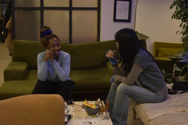 Two women are having a conversation in a bright ambient room with sofas and coffee table. The young dark-skinned woman on the left has her hair tied in a high bun. She is laughing and looking at the woman seated on the right whose profile is mainly obscured by her long. straight and dark hair.