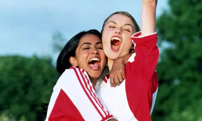 Two women in white and red football shirts hold onto each other while celebrating enthusiastically.