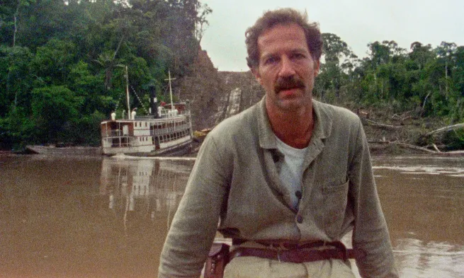 Werner Herzog faces the camera while behind him, on the other side of a murky river, a steamboat is being pulled up a muddy hil.