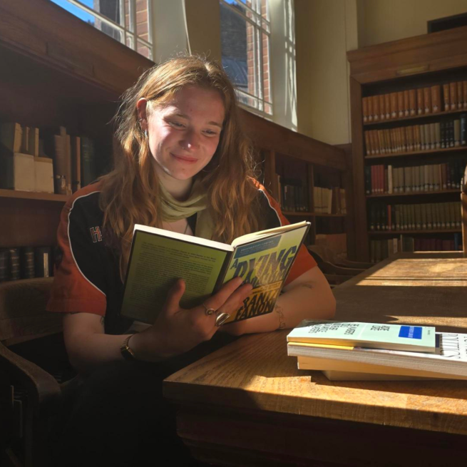 A woman with long ginger hair sits in a sunlit library reading a book