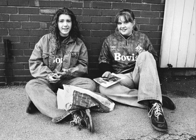 Two people sit on the ground against a brick wall. One wears a denim jacket and jeans; the other wears a sports jersey labeled “Bovis” and holds papers or magazines. A bag or package lies in front of them.