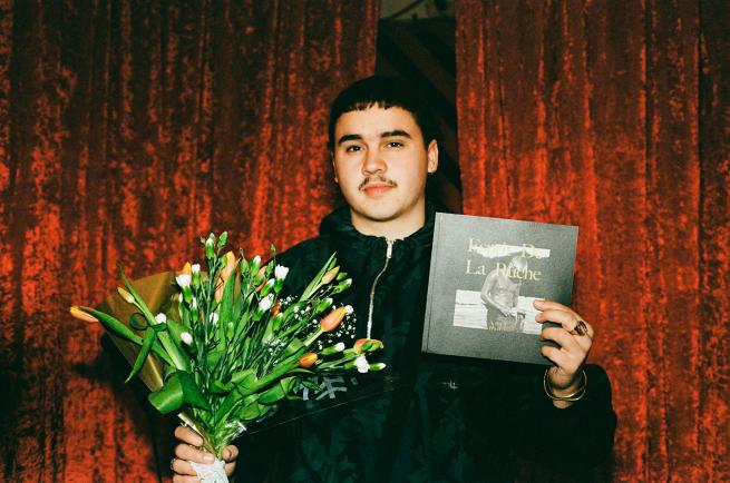 Image shows Aran hold a bunch of flowers and a book infornt of a red velvet curtain. 