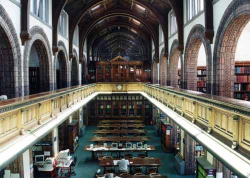Interior of a large historic library with high arched ceilings, wooden beams, and two levels. The upper level has a balcony with bookshelves; the lower level features long tables, computer workstations, and a person using a computer.