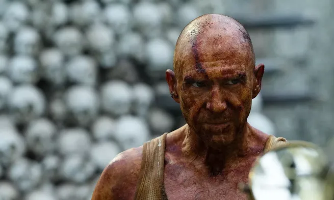 A man with a shaved head, wearing a dirty vest, and covered in blood and bruises