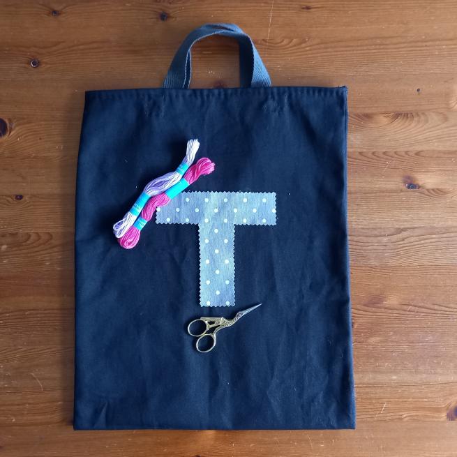 A blue tote bag which has a letter T laid on it in light blue fabric, there are two skeins of thread (light pink and dark pink) and a pair of embroidery scissors also on the tote bag.