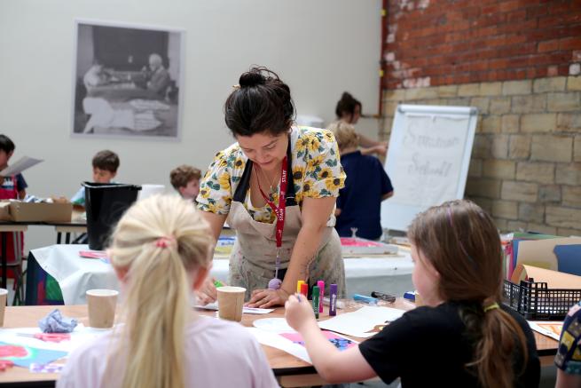 Image shows Gigi assisting at a workshops helping 2 young girls on screenprinting.