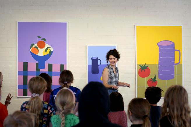 Image shows workshop facilitator Sonia taking to a group of young people. She standsinfornt of a wall which has 3 large still life images in bright and bold colours. 