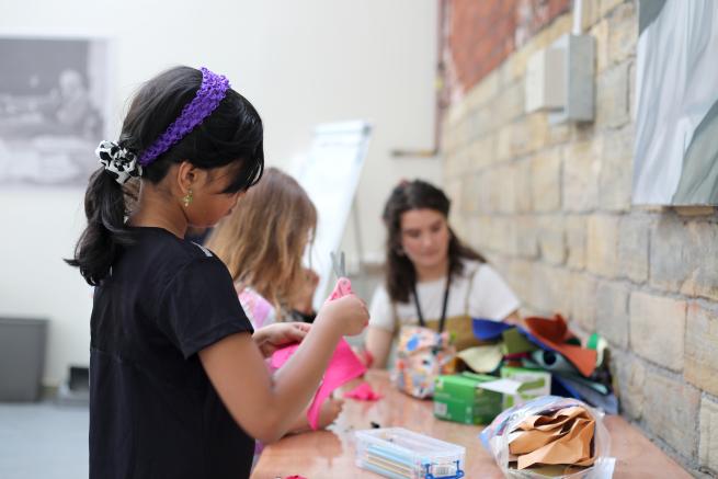 Image shows young girl working on a textile piece on a table, another young girl is in the background along with workshop facilitator Sonia. 