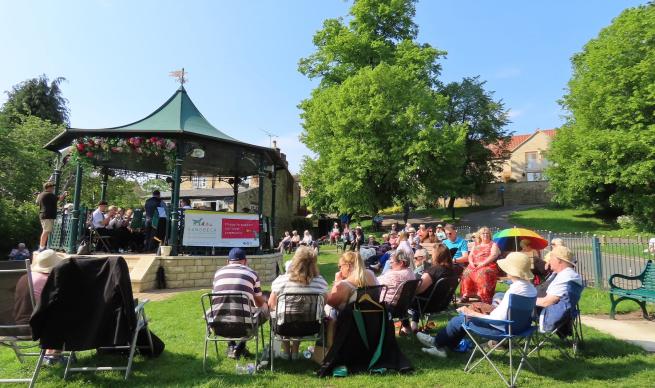 A sunny day with a crowd of people sat on deck chairs facing a band stand. There is a brass band performing on the bandstand.