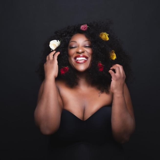 Judi Love from the waist up wearing a black top with her eyes closed, a big smile and flowers in her hair against a black background.