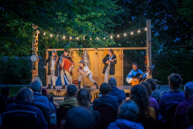 Night time in the forest. An enthralled audience watch actors on a wooden stage playing instruments. The scene is lit by festoon lights.