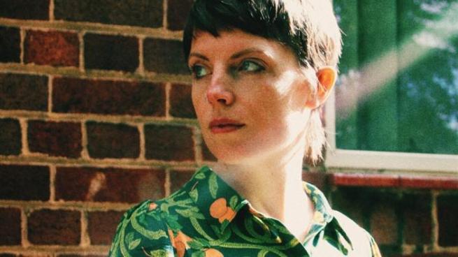 Quincey May Brown, has short brown hair and a floral green shirt, standing against a brick wall.