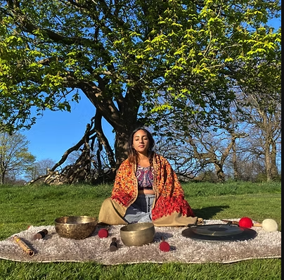 Neeta sat in a park, in front of trees, on a nice clear day,. She's sitting on a blanket, with various instruments of the sound bath. A gong, tibetan bowls, and bells. Her presence is calm and serene