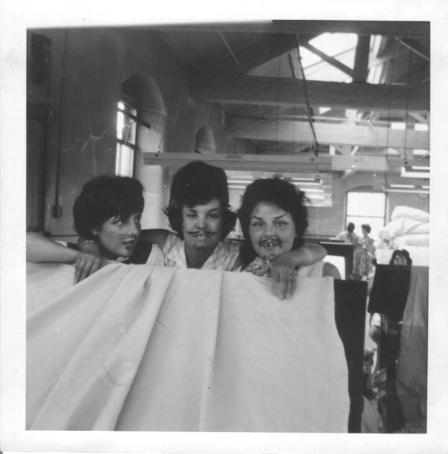 The black and white image shows three women in an old industrial space with drawn-on moustaches with a white sheet held infront of them. 
