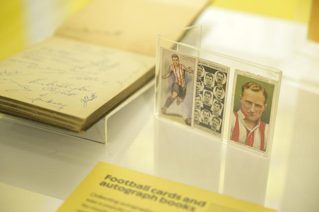 A display of football memorabilia featuring an open autograph book with multiple signatures on the left, and three football cards in a clear stand on the right. The cards include a player in action, a collage of headshots, and a blurred portrait. A yellow information card in front reads 'Football cards and autograph books.'