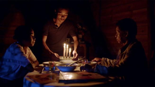 A young man puts a set of three lit candles on a table where two older men are sat in a dark room. 