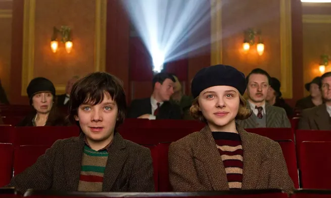 Hugo and his acolyte sit in the cinema, facing the camera. Adults sit behind them.