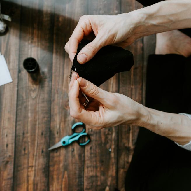 Two hands pinning a piece of black fabric in the foreground, blue sewing scissors lay on a wooden table in the background